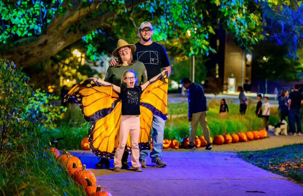 A family poses for a photo at the rain garden with a line of carved pumpkins featured in the background.